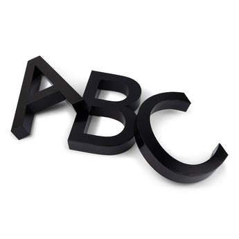 Drie grote, zwarte, 3D letters-A, B en C-Carlo Black 20cm by yessential.nl-liggen op een wit oppervlak, overlappen elkaar een beetje en staan onder verschillende hoeken als moderne huisnummers.