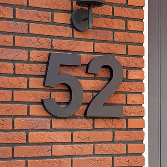 De Angelo Black 30cm huisnummers van yessential.nl zijn als moderne, grote, zwarte cijfers op een rode bakstenen muur gemonteerd onder een buitenlamp, met rechts het grijze deurkozijn zichtbaar voor een eigentijdse uitstraling.