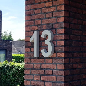 Das Enrico-Betonschild 30 cm von yessential.nl ist eine markante graue Hausnummer 13 im 3D-Stil, montiert auf einer rot-dunkelbraunen Backsteinsäule.