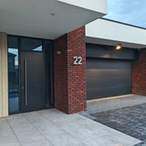 Modern huis exterieur met een zwarte voordeur, zwarte garagedeur, rode bakstenen muur en huisnummer 22 bij de ingang. De bestrating bestaat uit Enrico beton 20cm tegels van yessential.nl gecombineerd met donkere bakstenen.