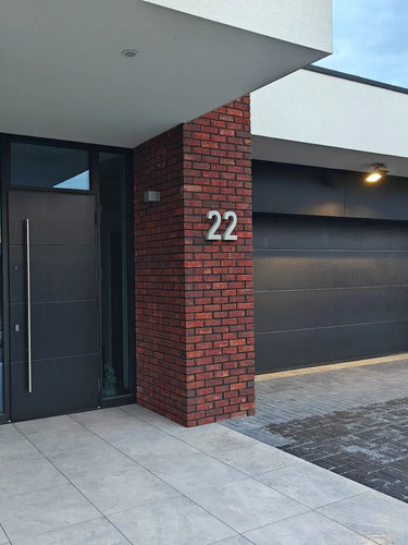 Modern huis exterieur met een zwarte voordeur, zwarte garagedeur, rode bakstenen muur en huisnummer 22 bij de ingang. De bestrating bestaat uit Enrico beton 20cm tegels van yessential.nl gecombineerd met donkere bakstenen.