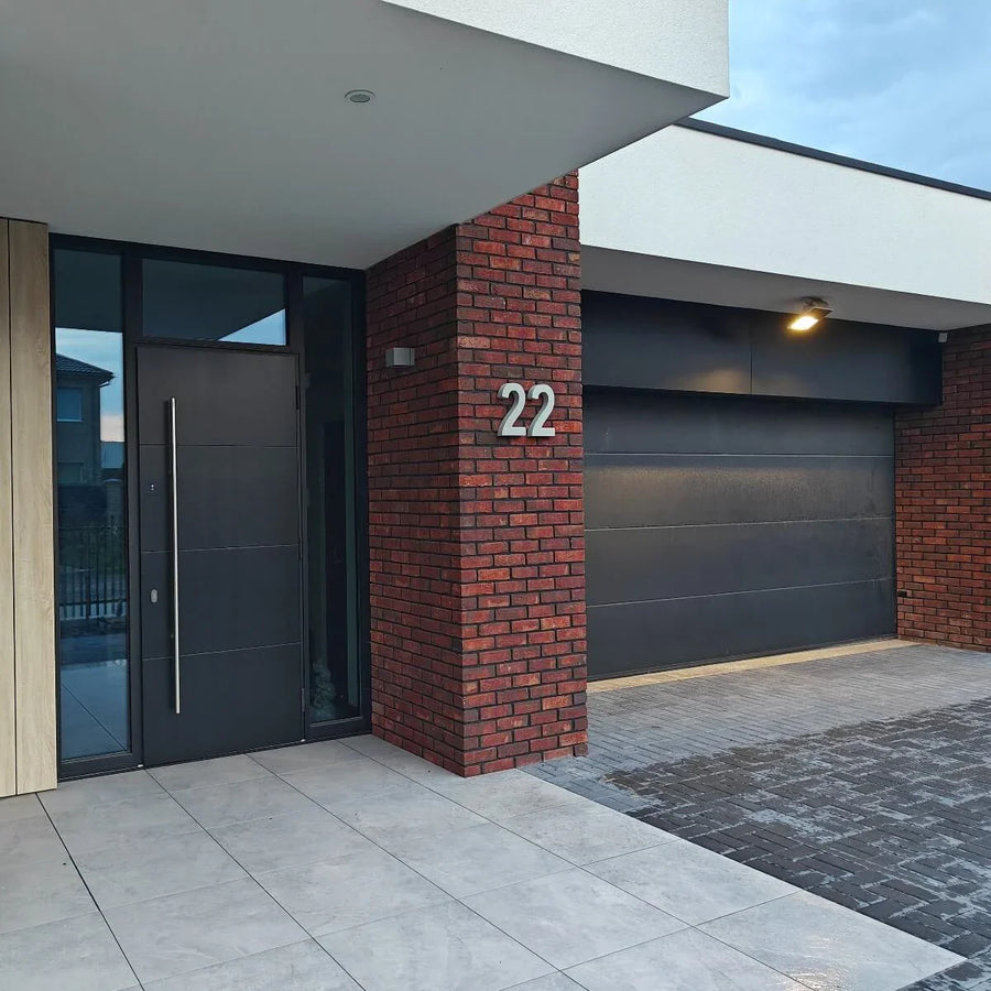 Modern huis exterieur met een zwarte voordeur, zwarte garagedeur, rode bakstenen muur en huisnummer 22 bij de ingang. De bestrating bestaat uit Enrico beton 20cm tegels van yessential.nl gecombineerd met donkere bakstenen.