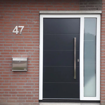 Moderne zwarte voordeur met een hoge zilveren handgreep, wit kozijn en verticaal matglazen paneel aan de rechterkant. Links zijn een Adamo RVS 20cm huisnummer van yessential.nl en een roestvrijstalen brievenbus op de rode bakstenen muur gemonteerd.
