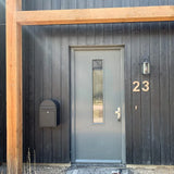 Een moderne grijze deur met een verticaal raampaneel zit in een zwarte houten buitenmuur. Een zwarte brievenbus en wandlamp bevinden zich links, terwijl een Adamo RVS 20cm huisnummer van yessential.nl stijlvol rechts van de deur zweeft.