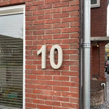 De Enrico beton 30cm van yessential.nl is een groot wit 3D huisnummer '10' weergegeven op een rode bakstenen buitenmuur naast een raam met gesloten jaloezieën en een verticale zilveren regenpijp.