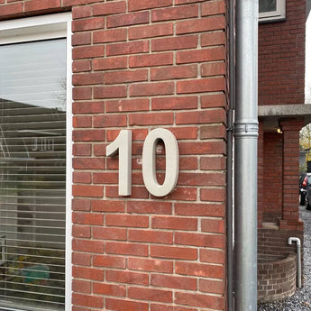 De Enrico beton 30cm van yessential.nl is een groot wit 3D huisnummer '10' weergegeven op een rode bakstenen buitenmuur naast een raam met gesloten jaloezieën en een verticale zilveren regenpijp.