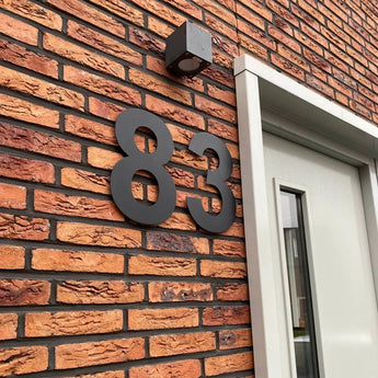 De Adamo Black 30cm huisnummers van yessential.nl zijn zwevend bevestigd op een bakstenen muur naast een moderne deur met glas en wit kozijn; boven de cijfers hangt een klein vierkant lichtarmatuur.
