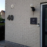 De buitenmuur van een licht bakstenen huis is voorzien van yessential.nl's Angelo Black 20cm huisnummer, een lamp boven de deurbel, een zwart bord met tekst en een deel van een glazen deur zichtbaar aan de rechterkant.