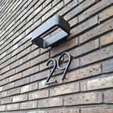 Het Amato Black 15cm huisnummer van yessential.nl is gemonteerd op een bruine bakstenen muur onder een rechthoekige zwarte buitenlamp en geeft de ingang van je huis een moderne stijl.
