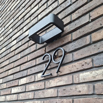 Het Amato Black 15cm huisnummer van yessential.nl is gemonteerd op een bruine bakstenen muur onder een rechthoekige zwarte buitenlamp en geeft de ingang van je huis een moderne stijl.