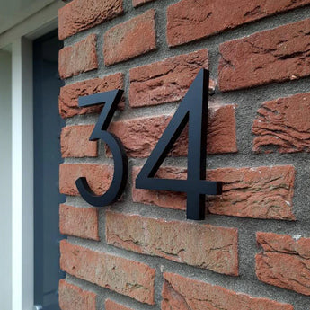 Milano Black 15cm huisnummers van yessential.nl, geïnstalleerd op een rode bakstenen muur bij een deur, creëren een moderne look doordat hun driedimensionale ontwerp gedurfde schaduwen werpt op de bakstenen.