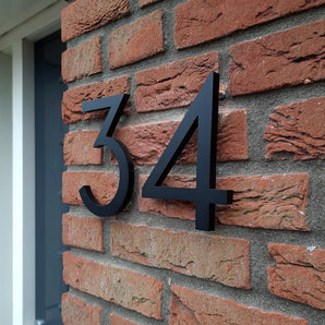 Milano Black 15cm huisnummers van yessential.nl, geïnstalleerd op een rode bakstenen muur bij een deur, creëren een moderne look doordat hun driedimensionale ontwerp gedurfde schaduwen werpt op de bakstenen.