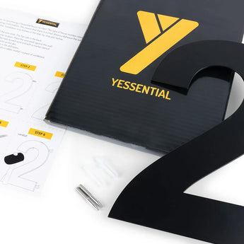 De Adamo Black 30cm van yessential.nl - een opvallend zwart metalen nummer 2 - bevat een instructieblad, installatiemateriaal en een zwarte doos met het gele Yessential-logo, allemaal netjes gepresenteerd op een wit oppervlak.