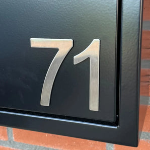 Close-up van Animo RVS 7,6cm (zonder boren) van yessential.nl, gemonteerd op een zwart structuur oppervlak met een bakstenen muur op de achtergrond - perfect als huisnummer zonder boren.