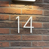 Het Amato White 12,5cm huisnummer van yessential.nl, gemonteerd op een rood en donkerbruine bakstenen muur met een rechthoekige lamp erboven, levert een moderne stijl - ideaal voor iedereen die op zoek is naar opvallende moderne huisnummers voor in huis.