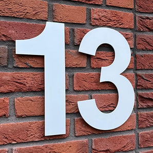 De Adamo White 30cm van yessential.nl is een groot wit nummer 13, ontworpen in de Adamo White stijl en gemonteerd op een rode bakstenen muur - een opvallend roestvrijstalen huisnummer voor een duidelijke adressering.