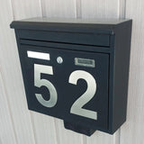 Een zwart metalen brievenbus met grote Animo RVS 15cm rvs huisnummers van yessential.nl is gemonteerd op een lichte, verticaal gepaneelde muur. De brievenbus heeft een postgleuf, sleutelgat en een rechthoekige naamplaathouder.