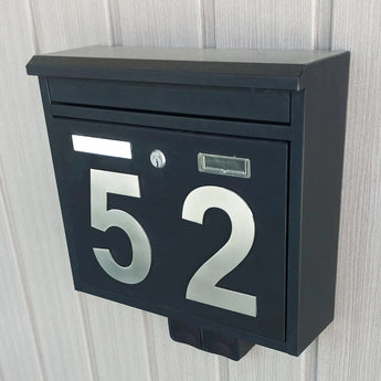Een zwart metalen brievenbus met grote Animo RVS 15cm rvs huisnummers van yessential.nl is gemonteerd op een lichte, verticaal gepaneelde muur. De brievenbus heeft een postgleuf, sleutelgat en een rechthoekige naamplaathouder.