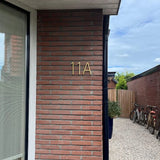 Een bakstenen buitenmuur is voorzien van Amato Gold 12,5cm huisnummers van yessential.nl. Op de achtergrond zorgen een grindpad, houten hek, potplanten en geparkeerde fietsen voor extra charme.