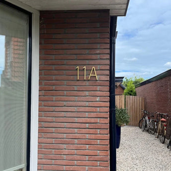 Een bakstenen buitenmuur is voorzien van Amato Gold 12,5cm huisnummers van yessential.nl. Op de achtergrond zorgen een grindpad, houten hek, potplanten en geparkeerde fietsen voor extra charme.