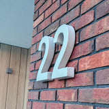 Het stijlvolle huisnummer Enrico beton 20cm van yessential.nl is duurzaam en gemonteerd op een rode bakstenen muur naast een deur met houten panelen en een vierkante lamp.