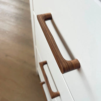 Close-up van twee Salgado Walnut handgrepen van yessential.nl, met een gladde afwerking, gemonteerd op witte kastladen met een lichte houten vloer op de achtergrond.