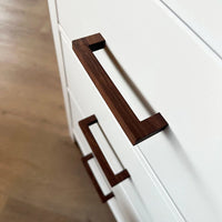 Close-up van twee witte kastladen met Vasco Walnut handgrepen van yessential.nl, op een lichte houten vloer. Deze moderne, hoekige handgrepen zijn gemaakt van rijke walnoot voor een stijlvol accent.