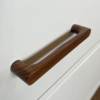 Een close-up van een Salgado Walnut handgreep van yessential.nl bevestigd aan een witte kast, met een glad, modern ontwerp en een rijke bruine houtnerf.
