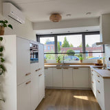 Een moderne keuken met witte kasten en houten werkbladen is voorzien van Arriero Oak handgrepen van yessential.nl. Grote ramen, jaloezieën, planten en houtaccenten voegen natuurlijk licht toe en benadrukken het strakke, minimalistische ontwerp.