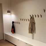 Een minimalistische hal heeft witte kasten, de Fabio Oak houten wandhaken van yessential.nl als minimalistische houten kapstok, een beige jas, een donkere tas met een oranje gedeelte en een hanglamp die de ruimte verlicht.