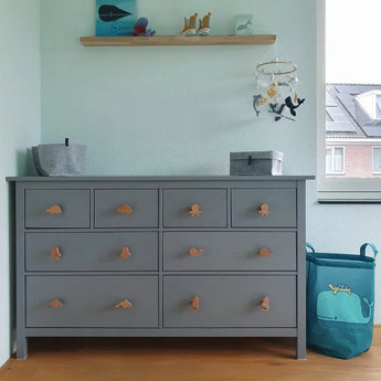 Een Bambino Dino grijze commode van yessential.nl staat tegen een lichtblauwe muur in de kinderkamer. Boven is een plank met decor, terwijl rechts een blauwe wasmand met een groen walvismotief staat. Een raam maakt het plaatje compleet.