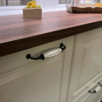 Een close-up van een keukenkastje met crèmekleurige deurtjes, een aanrecht van donker hout en de yessential.nl Daro Ceramic Black greep met zwarte details en een greep van wit keramiek. Op de achtergrond is een kleine plant op het aanrecht zichtbaar.