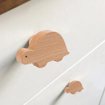 Een close-up van twee Bambino Dino houten ladeknoppen van yessential.nl, in de vorm van schildpadden en bevestigd op een licht kastje - eenvoudige, gladde accenten met stippenogen en natuurlijke houtnerf, perfect voor een kinderkamer.
