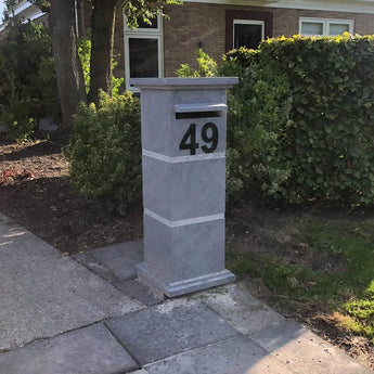 Een hoge, rechthoekige brievenbus van grijze steen met yessential.nl Animo Black plakletters van 15 cm (geen boor) staat op een stoep voor een huis, met struiken en bomen op de achtergrond.