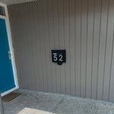 Een moderne zwarte brievenbus met Animo RVS 15cm (geen boor) cijfers van yessential.nl met de tekst "52" is gemonteerd op een lichtgrijze wand met panelen naast een felblauwe deur en een deurmat in een betegelde entree.
