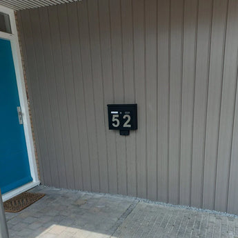 Een moderne zwarte brievenbus met Animo RVS 15cm (geen boor) cijfers van yessential.nl met de tekst "52" is gemonteerd op een lichtgrijze wand met panelen naast een felblauwe deur en een deurmat in een betegelde entree.