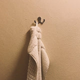 Een zachtbeige handdoek hangt gevouwen over het yessential.nl 20x Pagano Chroom haakje, dat is gemonteerd op een lichtbruine muur met structuur.