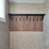 De 10x Avano Black wandkapstok van yessential.nl heeft een zwarte plank en stijlvolle haken op verticale houten latten, getoond in een neutrale kamer met een raam aan de linkerkant.