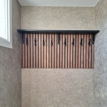 De 10x Avano Black wandkapstok van yessential.nl heeft een zwarte plank en stijlvolle haken op verticale houten latten, getoond in een neutrale kamer met een raam aan de linkerkant.