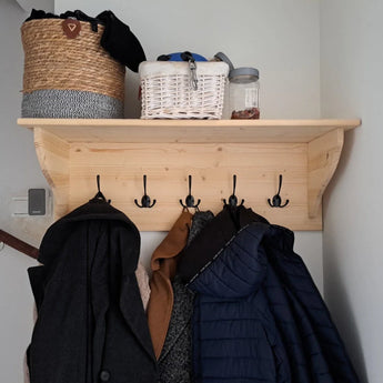 Een houten wandplank met vijf Nismo Black 2-haken van yessential.nl, waaraan jassen hangen. Op de plank staan een geweven mand, een witte mand, een grijze doek en een glazen pot - ideaal voor in de hal of entree.