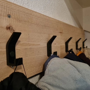 Een rij Huso Black haken van yessential.nl is gemonteerd op een houten plank met structuur en houdt meerdere jassen en jacks vast. De schuine opname benadrukt het strakke ontwerp van de haken tegen de houtnerf.