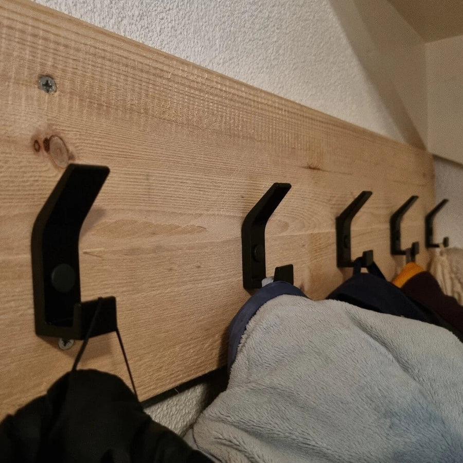 Een rij Huso Black haken van yessential.nl is gemonteerd op een houten plank met structuur en houdt meerdere jassen en jacks vast. De schuine opname benadrukt het strakke ontwerp van de haken tegen de houtnerf.