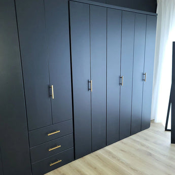 De Marano Gold kledingkast van yessential.nl heeft een modern donkergrijs ontwerp met goudkleurige minimalistische handgrepen, drie lades aan de linkerkant en meerdere hoge deuren, staande op een lichte houten vloer naast een wit gordijn.
