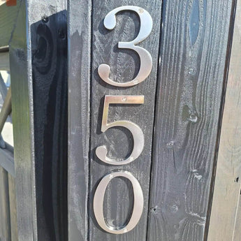 Matteo Nickel 10cm huisnummers van yessential.nl zijn verticaal gemonteerd op een donkere houten paal. Hun geborstelde metallic afwerking vangt zonlicht en werpt schaduwen die de textuur van het hout en de strakke glans van de nummers benadrukken.