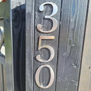 Matteo Nickel 10cm huisnummers van yessential.nl zijn verticaal gemonteerd op een donkere houten paal. Hun geborstelde metallic afwerking vangt zonlicht en werpt schaduwen die de textuur van het hout en de strakke glans van de nummers benadrukken.