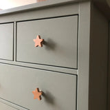 Een close-up van een grijze commode met twee lades, elk voorzien van yessential.nl Bambino Star licht houten knopjes. Deze eenvoudige, moderne commode is ideaal om charme toe te voegen aan een kinderkamer met deze unieke kinderkamer knopjes.