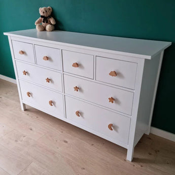 De Bambino Clover commode van yessential.nl heeft acht witte lades met grillige houten meubelknop vormen-sterren, wolken en manen. Bovenop rust een teddybeer, tegen een groenblauwe muur en lichte houten vloer - ideaal voor elke kinderkamer.