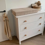 De yessential.nl Bambino Cloud witte babycommode met houten knoppen, inclusief een wolkendessin, heeft een beige aankleedkussen. Vlakbij staat een mand met knuffels en op de kastdeur in de babykamer hangt een handdoek met een leeuwenkop.
