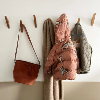 Een bruine leren handtas, beige sjaal, roze cartoon pufferjack en groene gewatteerde jas hangen op de yessential.nl Fabio Oak kapstok tegen een tweekleurige witte en beige muur.