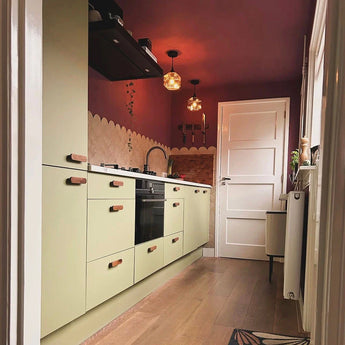 Een moderne kombuiskeuken met lichtgroene kastjes, yessential.nl Arriero Walnut handgrepen, houten vloeren, geschulpte beige achterwand, dieprode muren en plafond, hanglampen en een witte deur aan het einde. Natuurlijk licht stroomt binnen via een raam aan de rechterkant.